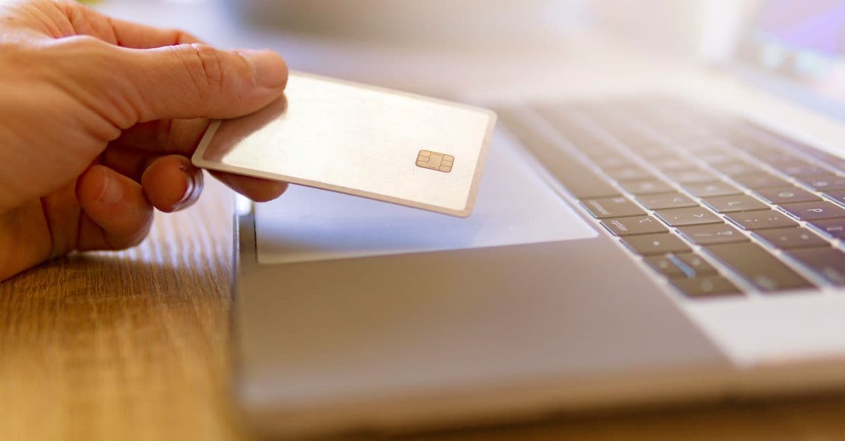 Person som betaler faktura med kredittkort på mobil og laptop ved kjøkkenbordet